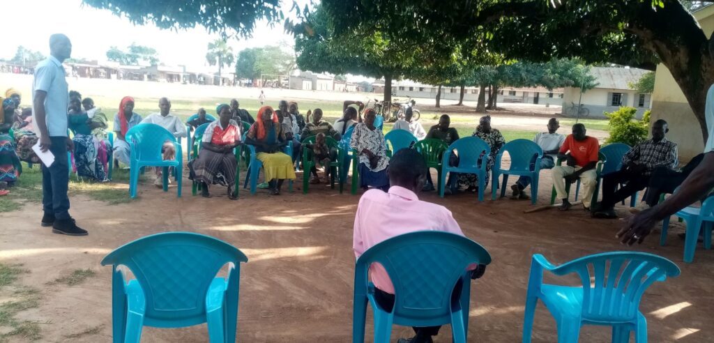 David facilitating a community dialogue meeting on C4D with local residents seated outdoors in Budaka District, Uganda